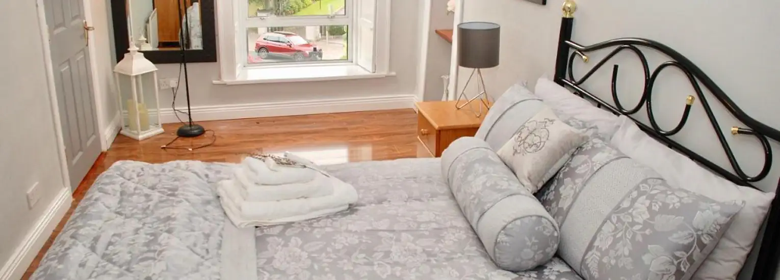 A bright bedroom with a neatly made bed in grey floral bedding, folded towels on top, a wooden bedside table with a lamp, and a large window overlooking a street with a red car parked outside.