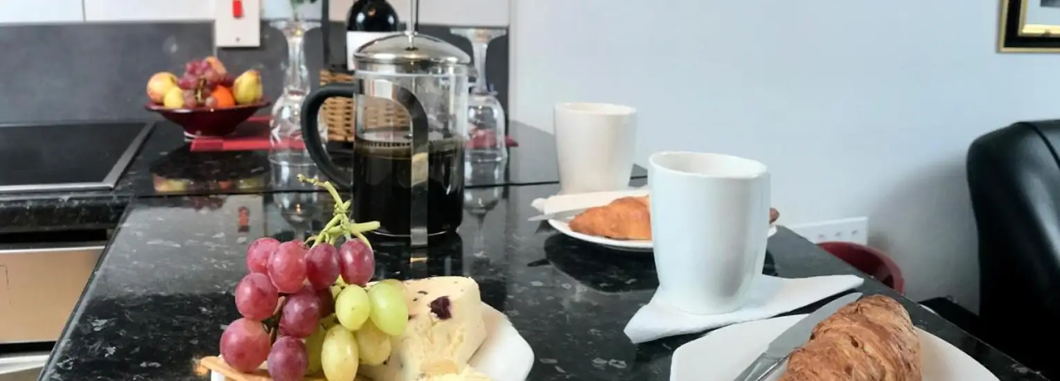 A kitchen worktop with a plate of red and green grapes, cheese, a croissant, two white mugs, a cafetière with coffee, and a bowl of fruit in the background.