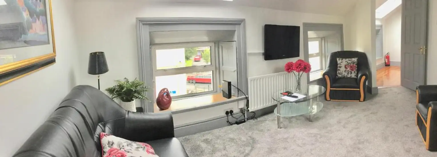 A cosy living room with grey carpet, black leather sofas, floral cushions, a glass coffee table with pink flowers, a wall-mounted TV, a radiator, two small windows, and modern decor.