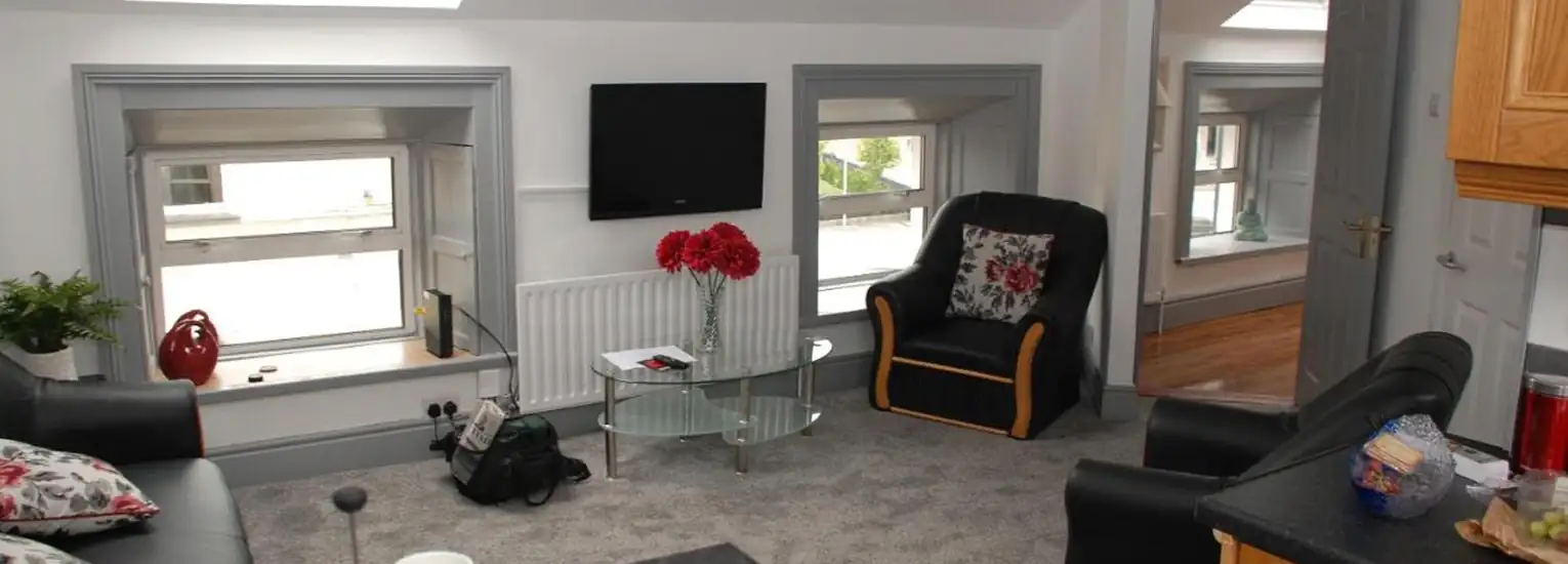 A cosy living room with grey carpet, black sofas with floral cushions, a glass coffee table with red flowers, a TV mounted on the wall, and two large windows. An armchair and adjoining room are visible.