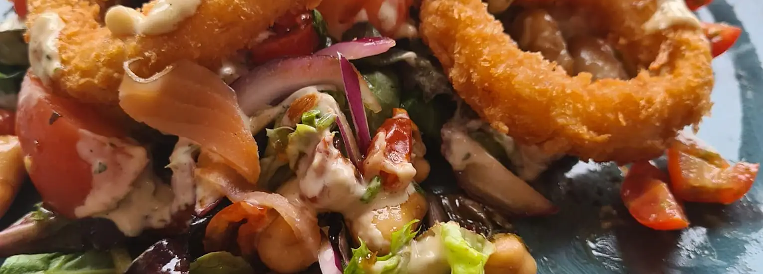 Close-up of a colourful salad topped with crispy fried onion rings, red onion slices, cherry tomatoes, chickpeas, mixed leaves, and a creamy dressing.