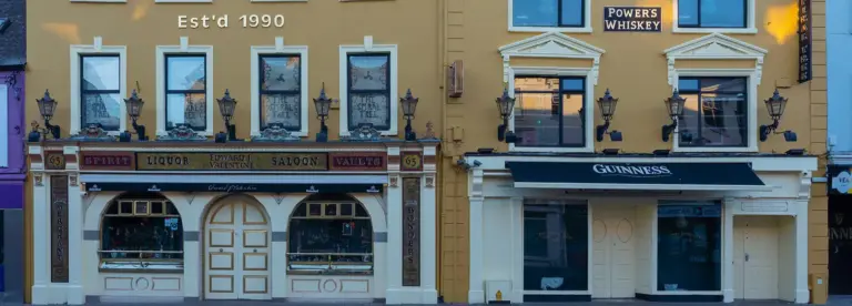 Two adjacent pubs with yellow façades, vintage lamps, and signage for Guinness and Powers Whisky. The left pub has arched windows and doors, with "Est'd 1990" above. Both buildings have ornate details and closed windows.