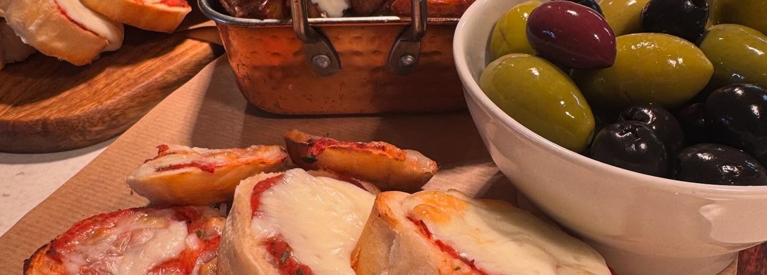 A close-up of a spread featuring slices of bread with melted cheese and tomato sauce, a bowl of assorted green and black olives, and a small copper basket of roasted potatoes in the background.