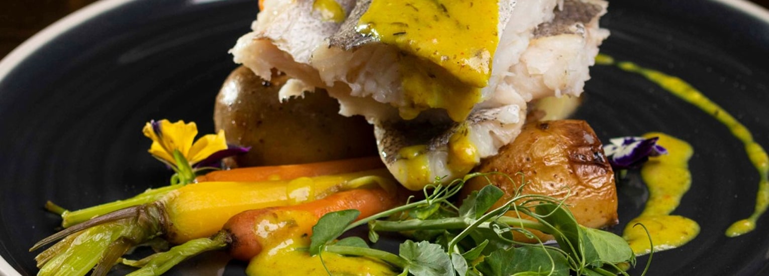 A gourmet dish featuring a fillet of white fish topped with yellow sauce, served with roast potatoes, baby carrots, greens, and edible flowers on a dark plate.