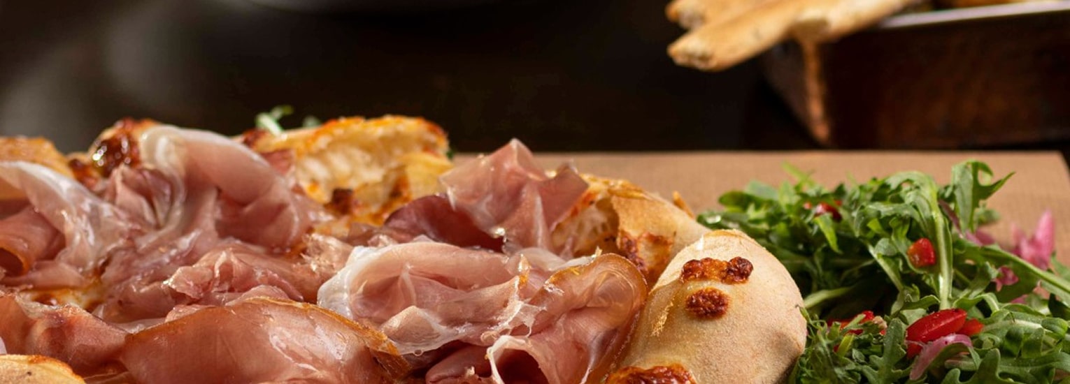 A close-up of a pizza topped with thin slices of prosciutto, next to a fresh green salad with rocket and red pomegranate seeds. Breadsticks are visible in the background.