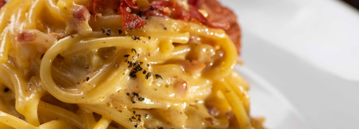 Close-up of creamy spaghetti carbonara topped with crispy bacon bits and freshly ground black pepper on a white plate.