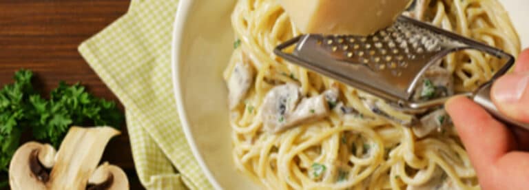 A close-up of creamy mushroom pasta in a white bowl, with someone grating cheese over the top. Parsley and sliced mushrooms are on the side, next to a green chequered serviette.