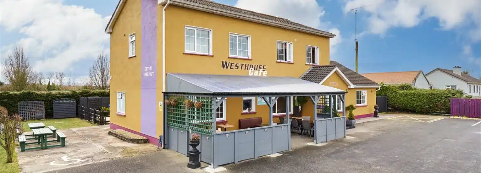 A yellow two-storey building with "Westhouse Cafe" signage, an outdoor seating area under a grey canopy, picnic tables on the grass, and a car park in front, set on a clear day.