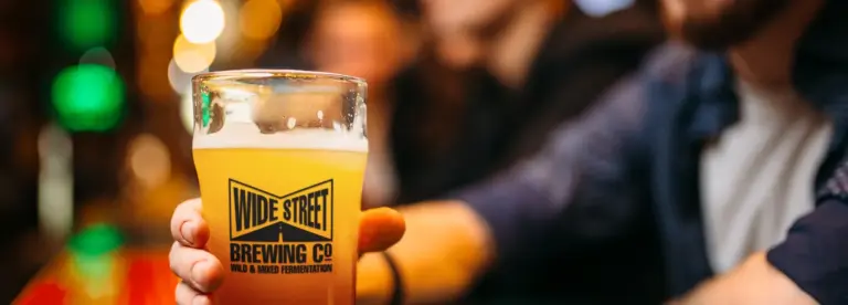 A person holds a pint glass of hazy beer with a "Wide Street Brewing Co." logo at a bar; the background is softly blurred with warm lighting and other people present.