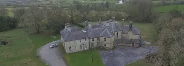 A large, old manor with multiple chimneys is surrounded by trees and green fields. A curved drive leads to the house, with a car parked nearby. The landscape is rural and expansive.