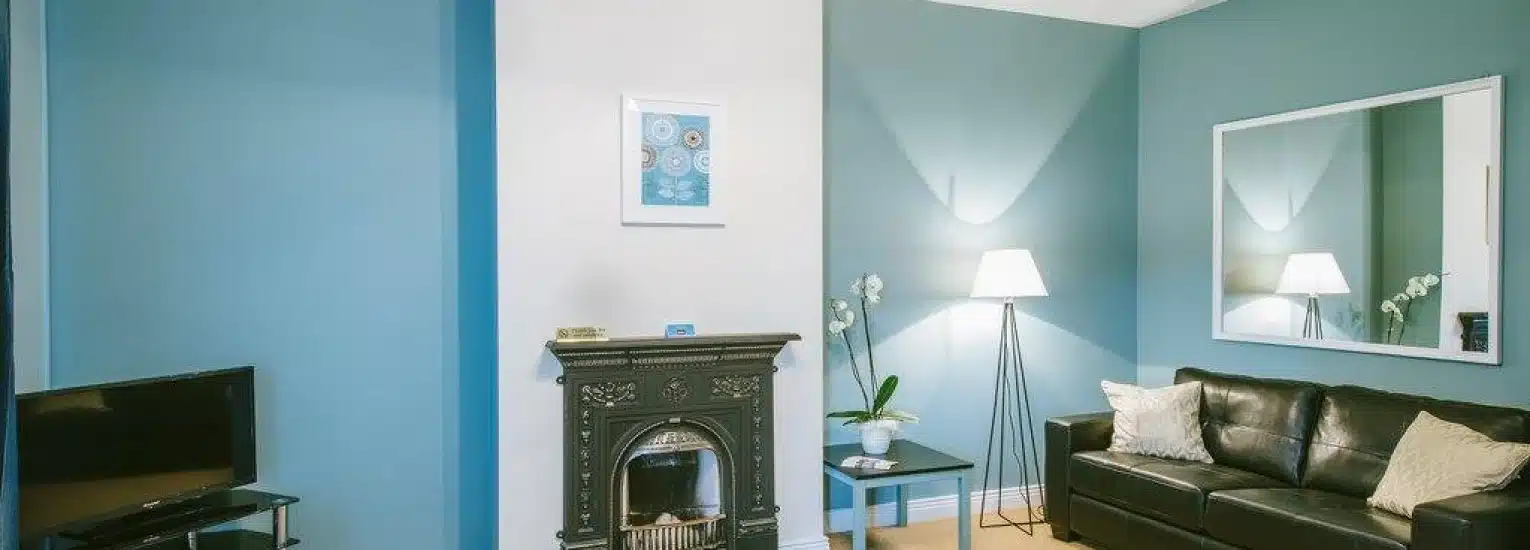 A modern living room with blue and white walls, a black sofa with cream cushions, a standard lamp, a fireplace, a large wall mirror, a television, and a small glass table with a potted plant.