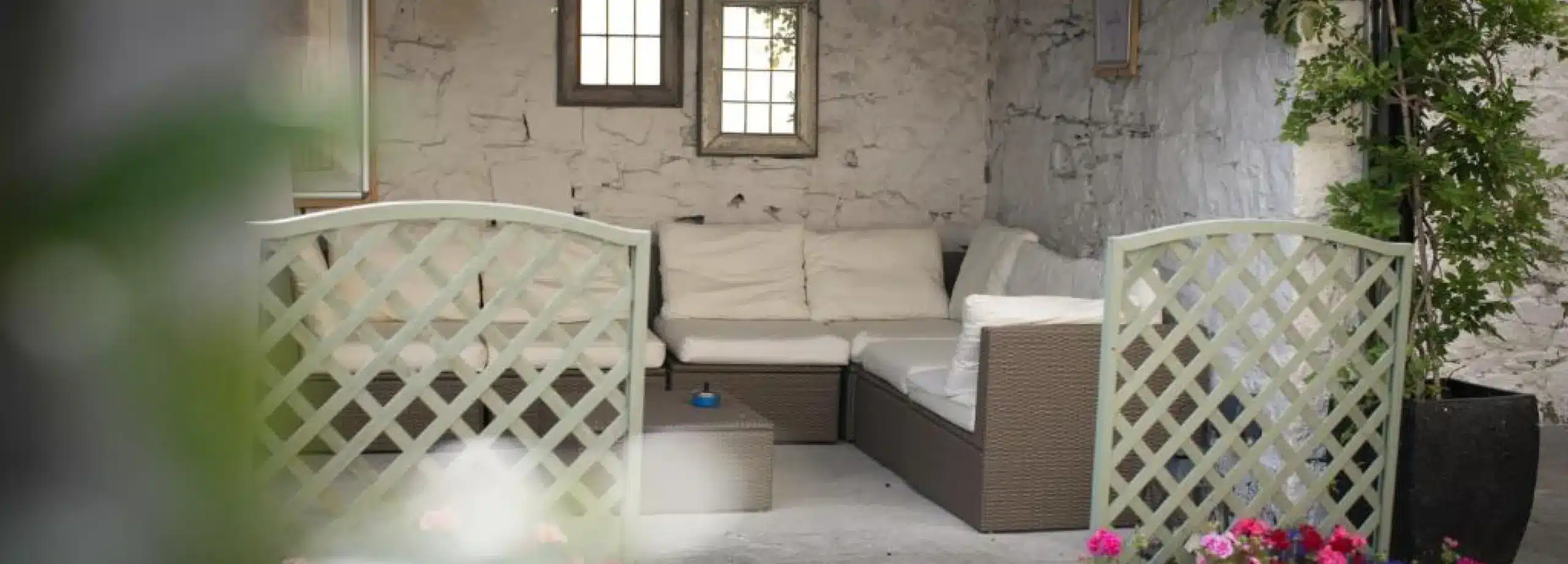 Outdoor seating area with cushioned wicker sofas set against a white stone wall, two small windows above, and lattice panels in front. Pink flowers and greenery are visible in the foreground.