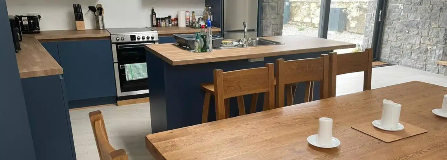 Modern kitchen and dining area with wooden worktops, navy blue cupboards, a cooker, and wooden dining table with chairs. Three white pillar candles are on the dining table. Large windows let in natural light.