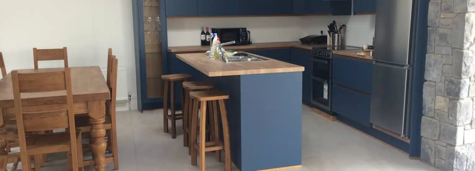 Modern kitchen with blue cupboards, a wooden dining table with six chairs, an island with stools, stainless steel appliances, and stone accents on one wall. Bottles and kitchenware are visible on the worktops.