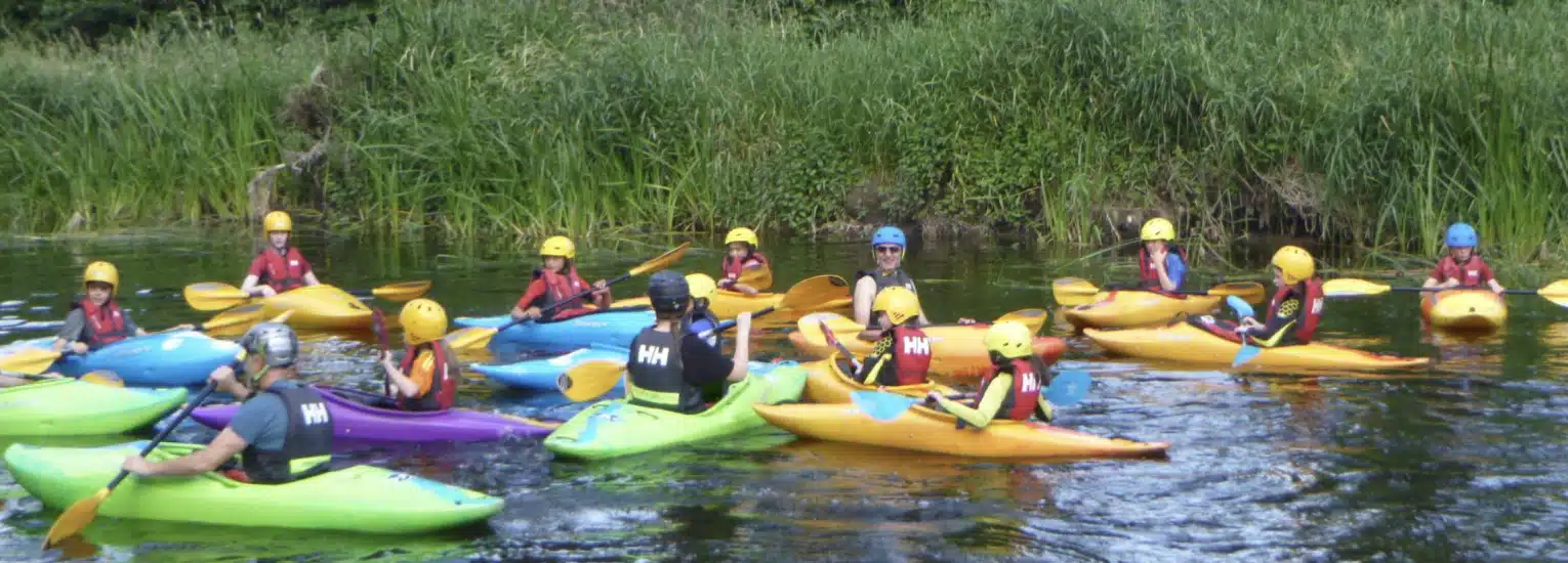 A group of people wearing helmets and life jackets paddle colourful kayaks on a calm river surrounded by tall green grass.