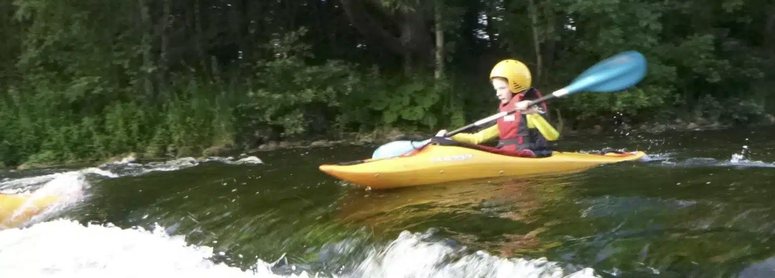 A person wearing a yellow helmet and life jacket paddles a yellow kayak on a river with small waves, with green trees in the background.
