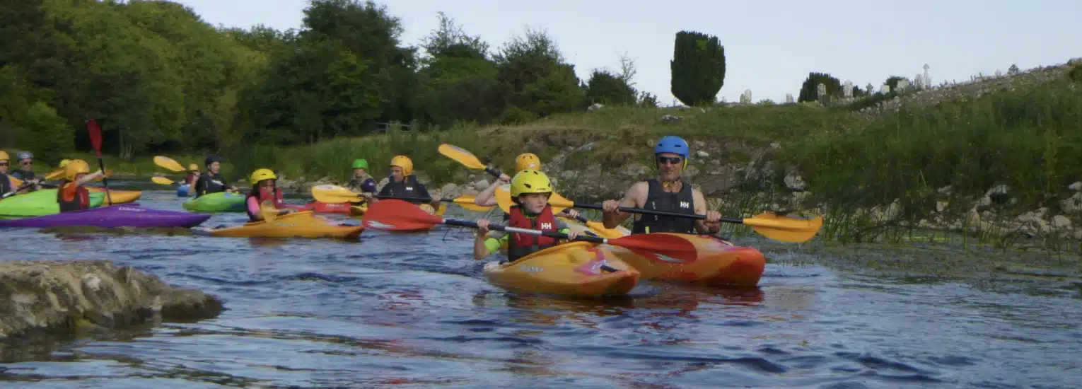 A group of people, mostly children, wearing helmets and life jackets kayak together on a river surrounded by greenery on a sunny day.