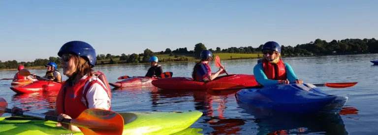 Several people wearing helmets and life jackets paddle colourful kayaks on a calm lake, with trees and grassy hills visible in the background under a clear blue sky.