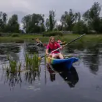 A person in a red shirt paddles a blue kayak on a calm river, with another kayak and greenery in the background under a cloudy sky.