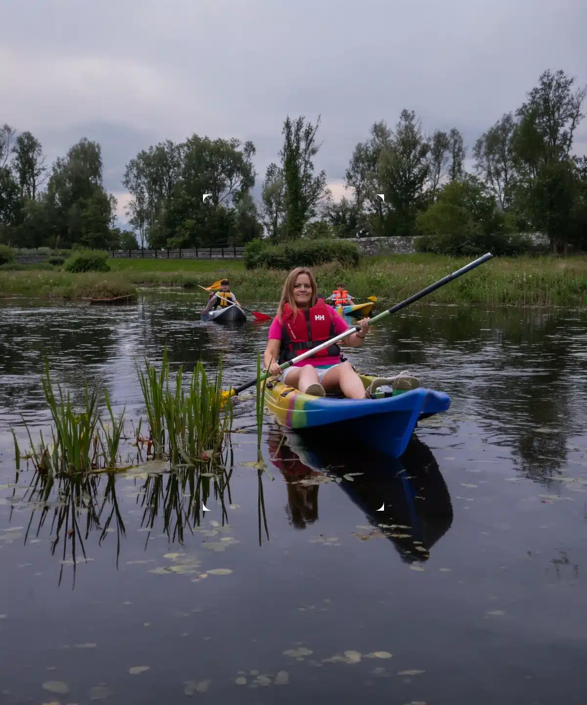 A person in a red shirt paddles a blue kayak on a calm river, with another kayak and greenery in the background under a cloudy sky.