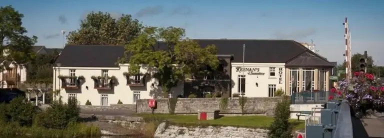 A cream-coloured building with the sign "Keenan's Hotel" sits beside a stone wall and river, surrounded by trees and flowers under a clear blue sky—an inviting spot to Visit Longford.