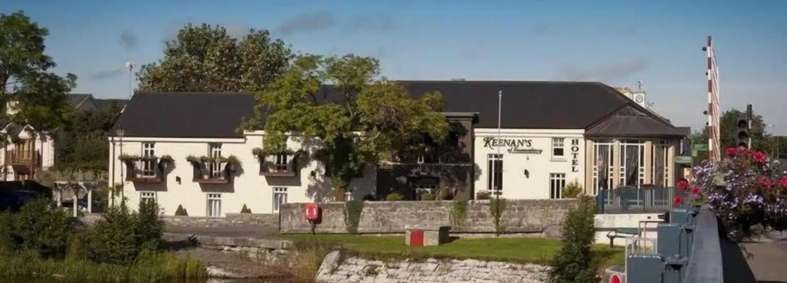 A cream-coloured building with the sign "Keenan's Hotel" sits beside a stone wall and river, surrounded by trees and flowers under a clear blue sky—an inviting spot to Visit Longford.