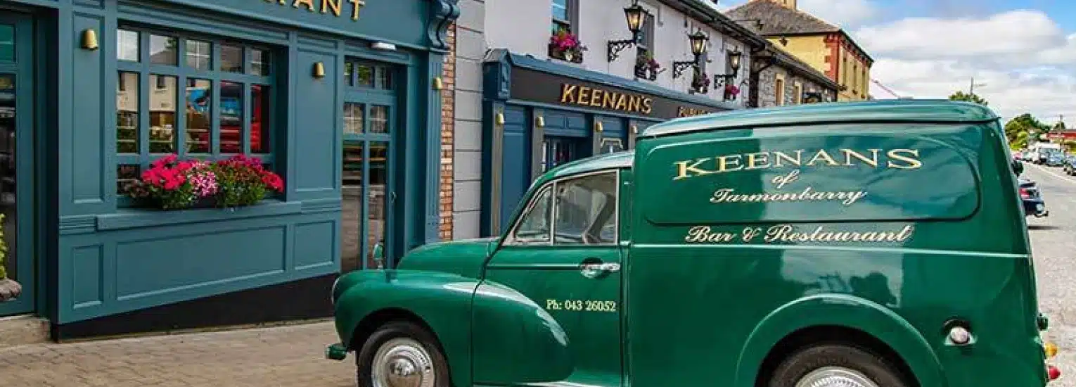 A vintage green delivery van labelled "Keenans of Tarmonbarry Bar & Restaurant" is parked outside a blue-painted pub with flower boxes beneath its windows on a sunny day.
