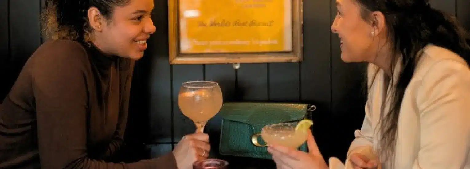 Two women sit opposite each other at a table in a restaurant, smiling and holding cocktails, engaged in conversation. A green handbag rests on the table between them.