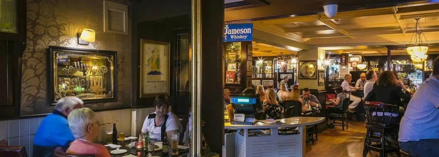 People dining and socialising inside a cosy, warmly-lit pub decorated with framed pictures and memorabilia on the walls. Tables are filled with guests, and a Jameson Whisky sign hangs above the counter.