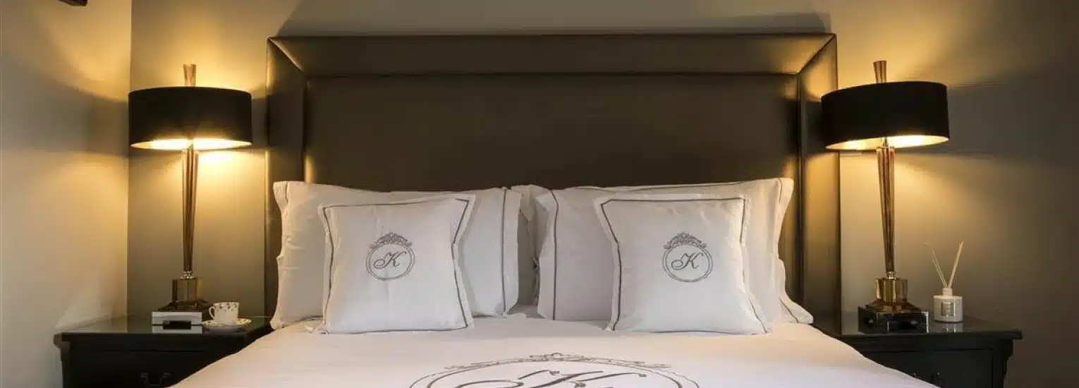 A neatly made bed with crisp white linen and decorative cushions sits between two black bedside tables, each holding a modern black and gold lamp. The headboard is upholstered in dark material.