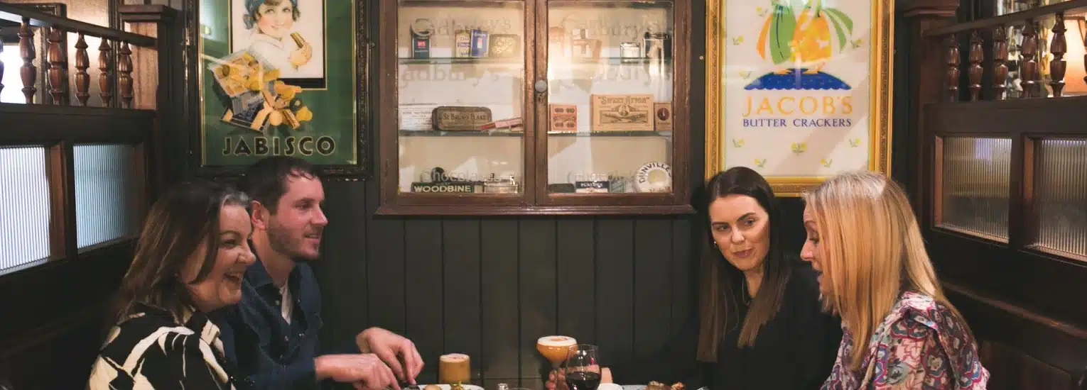Four people sit around a table in a cosy café, enjoying drinks and chatting. Vintage posters and shelves with glass jars decorate the walls behind them.