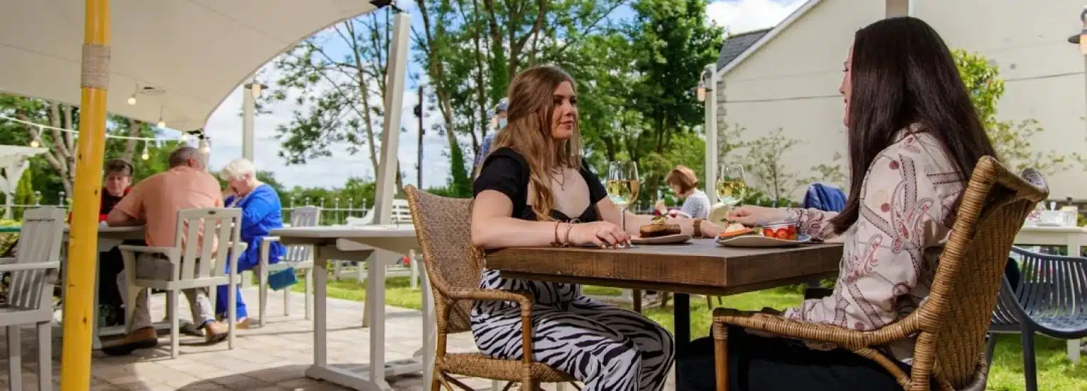 Two women sit at an outdoor restaurant table with food and drinks, talking. Other people are seated at tables in the background under a canopy, with trees and buildings around. It is a sunny day.