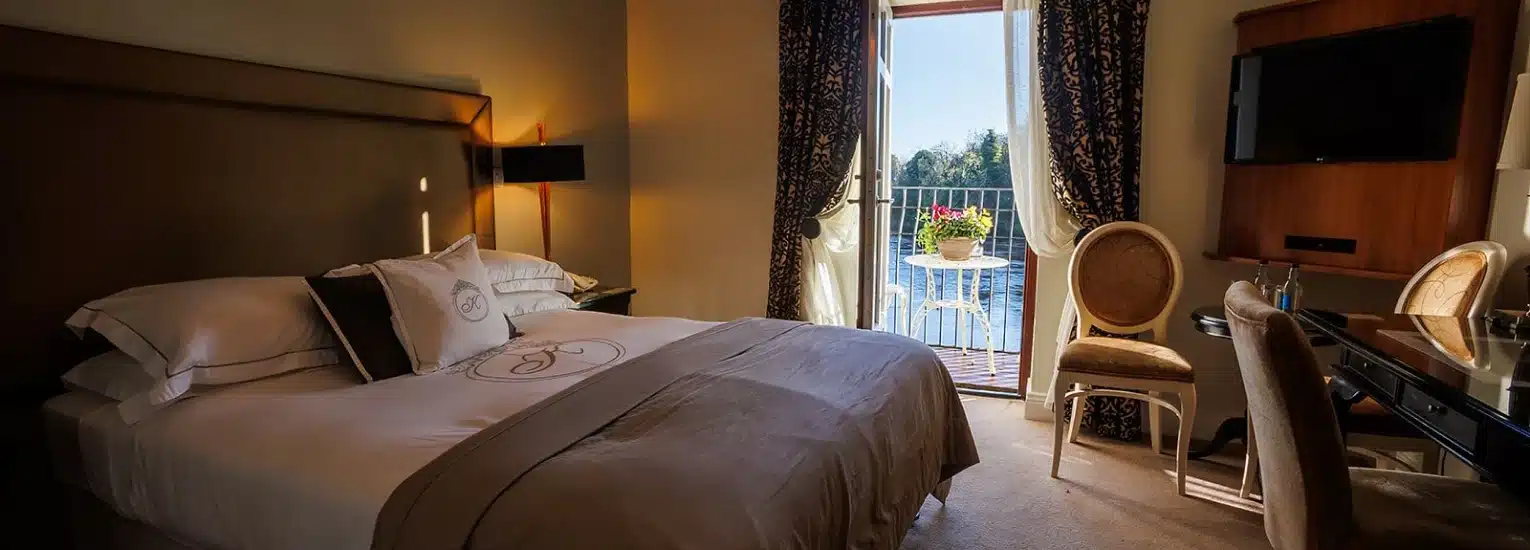 A cosy hotel room with a large bed, elegant chairs, a desk, and a wall-mounted TV. Sunlight streams in through a glass door leading to a balcony with a view of water and trees outside.