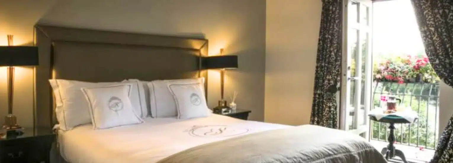 A cosy bedroom with a large bed, two bedside lamps, patterned curtains, and an open door leading to a balcony with flowers and outdoor furniture. Natural light brightens the room.