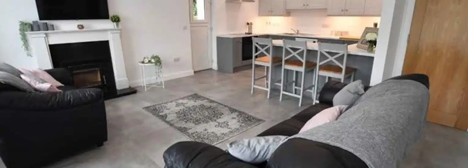 Modern open-plan living room and kitchen with black sofas, a grey rug, a fireplace, and a counter with white bar stools. The space features light-coloured walls, tiled flooring, and wooden accents.