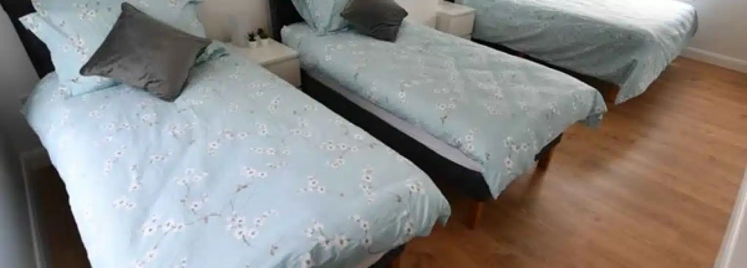 Three single beds with light blue floral bedding are arranged side by side in a room with wooden flooring. Each bed has a pillow and a small white bedside table positioned between them.