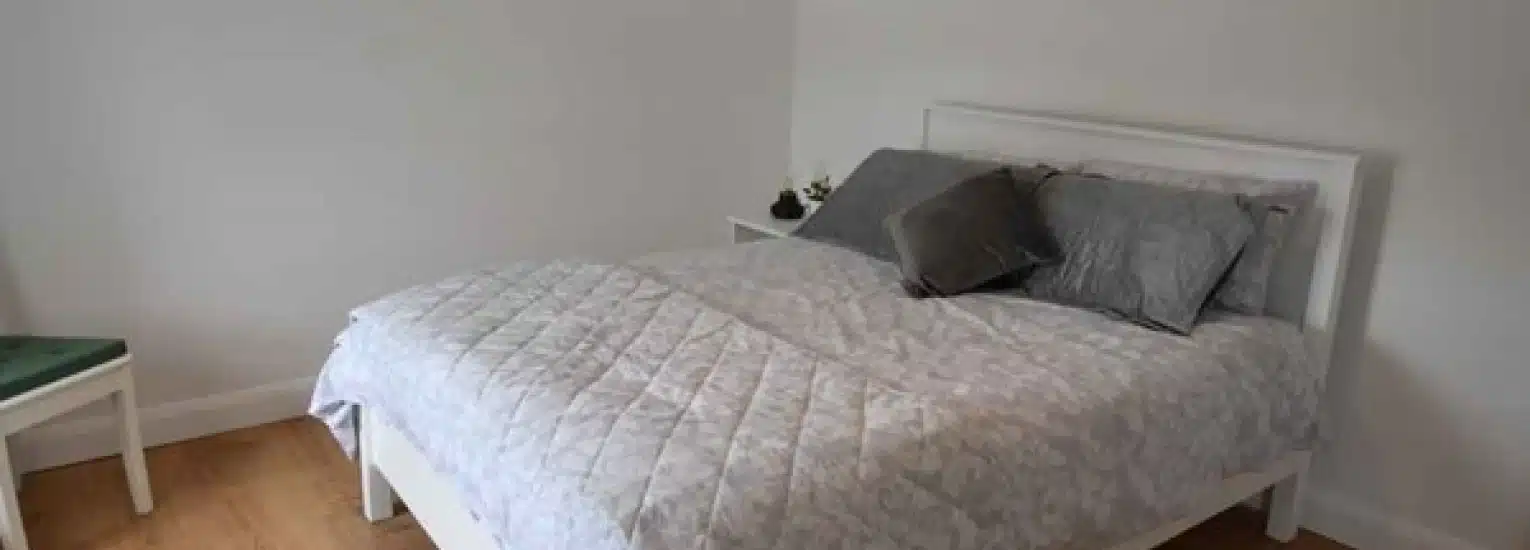 A neatly made bed with a white frame, light grey patterned bedding, and grey pillows in a simple, minimalistic bedroom with white walls and wooden floor. A small green chair and a bedside table are visible.