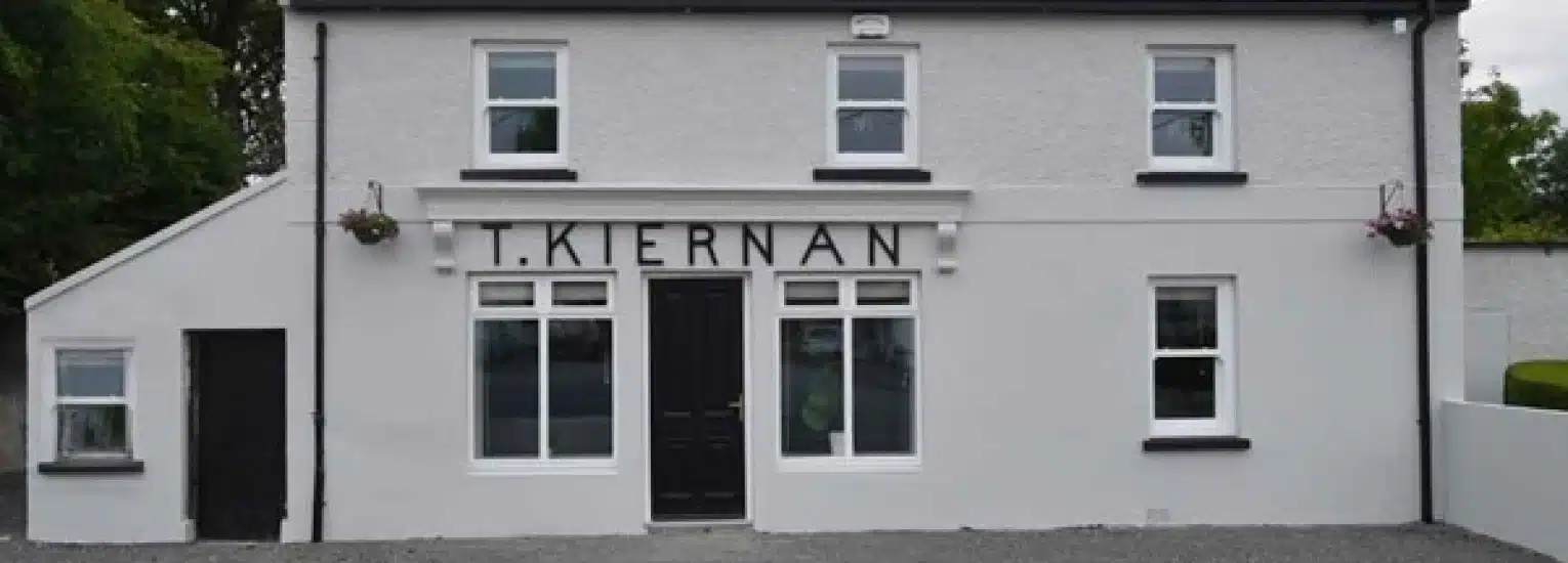 A white two-storey building with black trim, featuring a central black door and six windows. Above the door is a sign reading "T. KIERNAN." Two hanging flower baskets are on either side of the building.