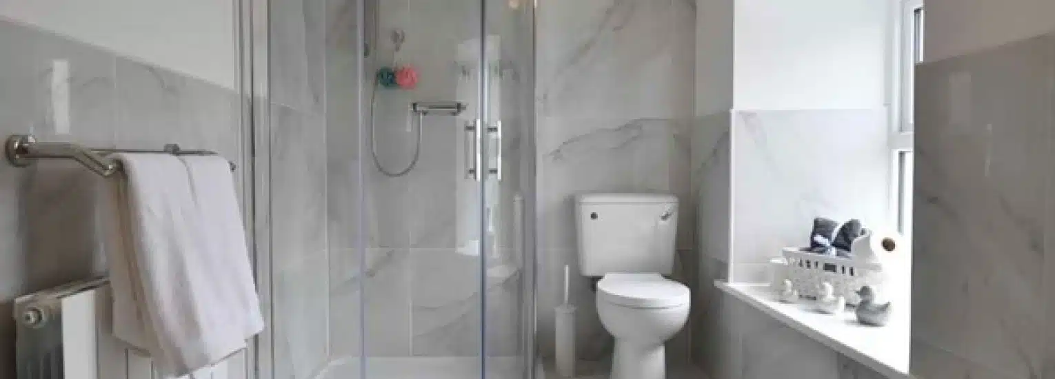 A modern bathroom with grey marble walls, a glass-enclosed shower, a white toilet, a towel on a heated rail, and a window sill with decorative items and towels.