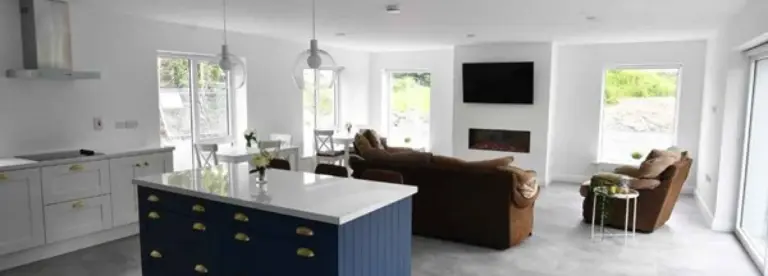 Modern open-plan living room and kitchen with white walls, large windows, blue kitchen island, brown sofas facing a wall-mounted TV above a fireplace, and light grey flooring.