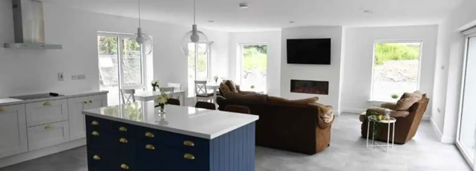 Modern open-plan living room and kitchen with white walls, large windows, blue kitchen island, brown sofas facing a wall-mounted TV above a fireplace, and light grey flooring.