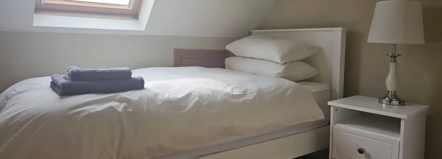A neatly made single bed with white bedding and pillows, a stack of folded grey towels on top, next to a white bedside table with a lamp under a skylight window in a softly lit room.