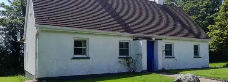 A small white cottage with a dark brown gabled roof, blue door, and four windows sits amid green grass and trees under a clear sky—an inviting sight for those who visit Longford.