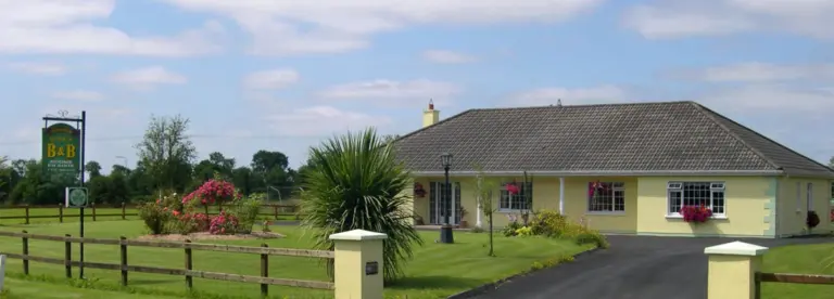 A single-storey yellow bed and breakfast building with a grey tiled roof, surrounded by a well-kept lawn, flowers, a driveway, and a wooden fence; a signpost for the B&B stands at the entrance.