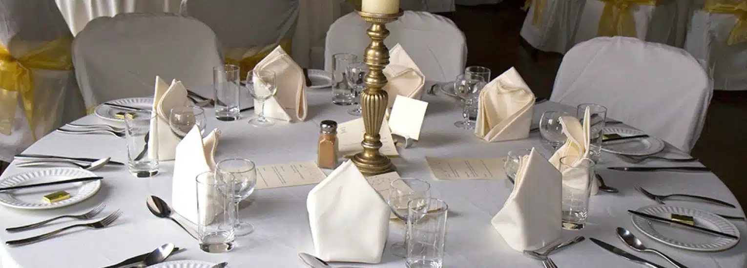 A round table set for a formal event with white tablecloth, neatly folded white serviettes, glassware, cutlery, plates, a menu, and a gold candle holder centrepiece. White chairs with yellow sashes surround the table.