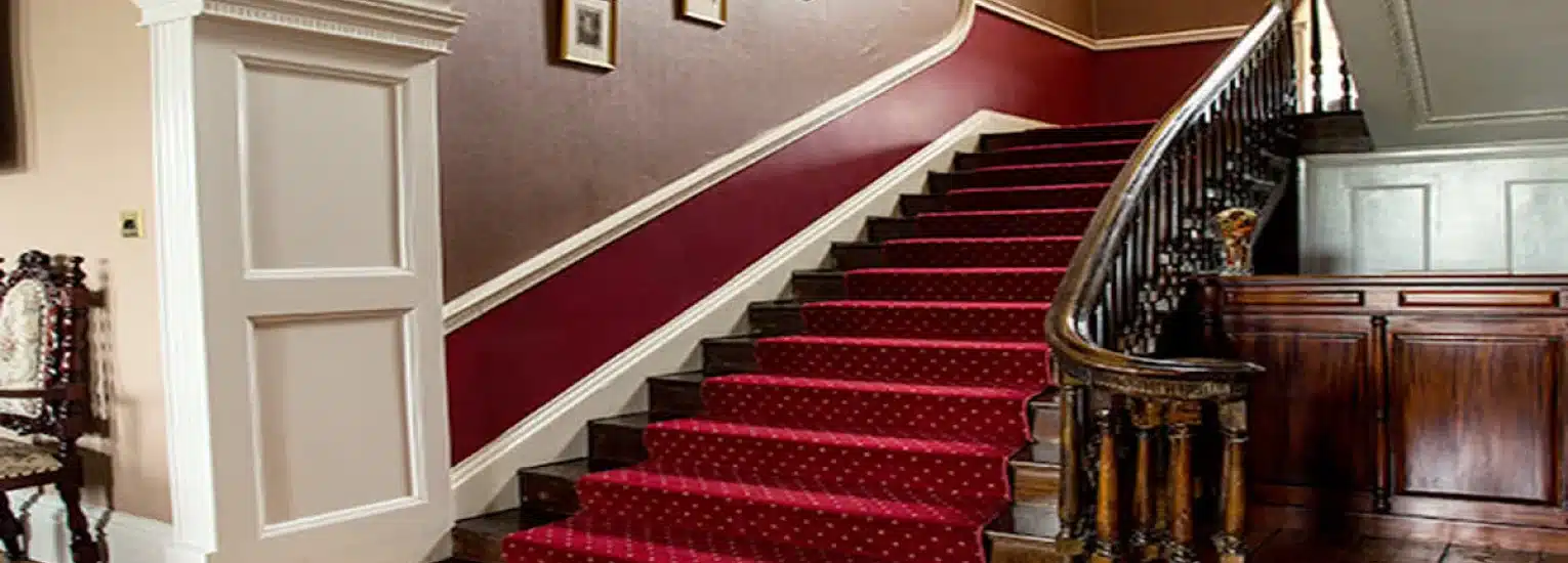 Curved staircase with a red carpet and wooden banister, set against red and beige walls. Ornate wooden furniture and framed pictures decorate the hall.