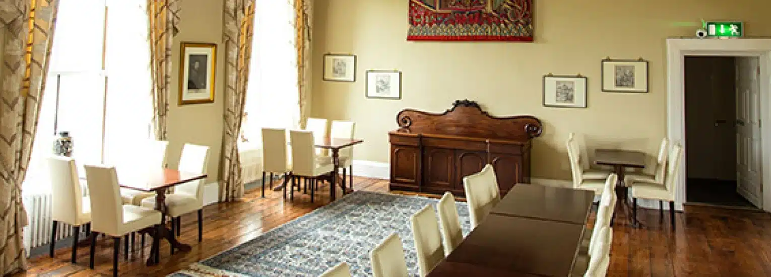 A bright, elegant room with wooden floors, cream walls, white chairs, dark tables, large windows with patterned curtains, framed art on the walls, and a patterned rug on the floor.