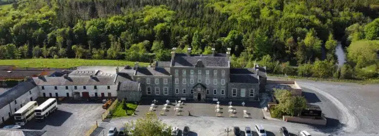 A large historic stone building with a central courtyard, surrounded by trees and greenery. Outdoor tables and chairs are arranged in front, with parked vehicles nearby and a hillside forest in the background.