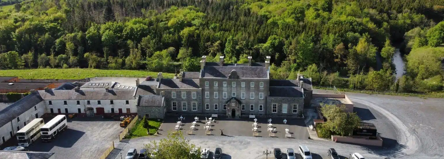 A large historic stone building with a central courtyard, surrounded by trees and greenery. Outdoor tables and chairs are arranged in front, with parked vehicles nearby and a hillside forest in the background.