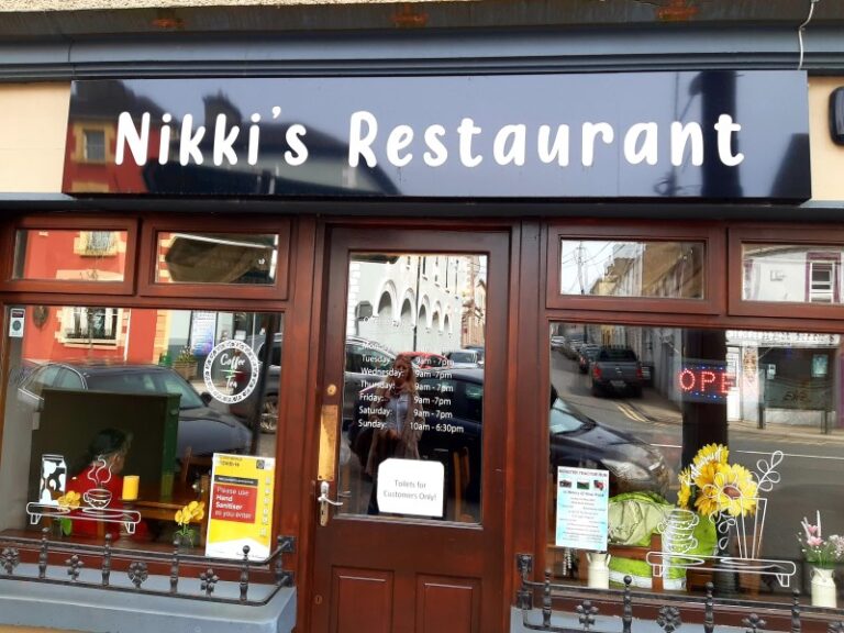 The exterior of Nikki’s Restaurant with large windows, a dark blue sign with white lettering, visible flowers and decorations inside, and an "OPEN" sign on the door. Reflections of nearby buildings appear in the glass.
